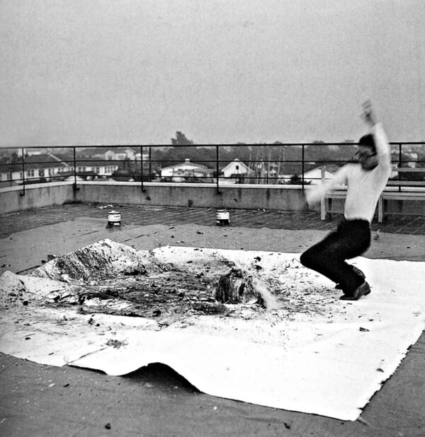 Black and white photograph of a person standing in an active position on a large white square covered in objects and splattered liquid