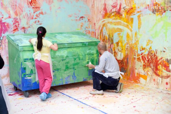 Two people paint a dumpster green in a space with colorful, haphazardly painted walls in pinks, blues, reds, and yellows