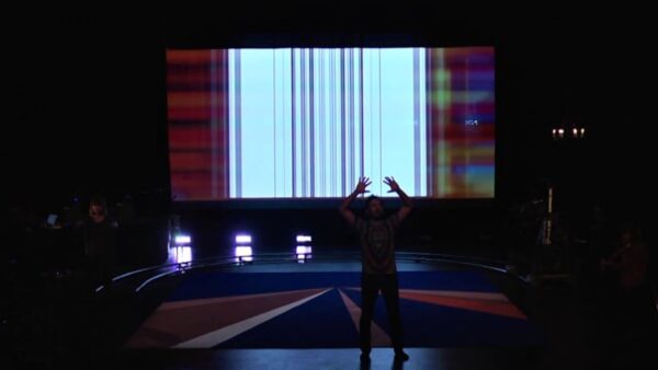 A person raises their hands above their head in a sign of submission on a dark stage illuminated from behind by a large screen featuring vertical bars of static