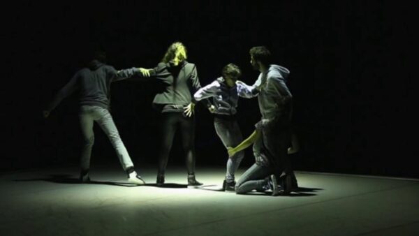 Four people hold onto each other on a dark stage with one spotlight