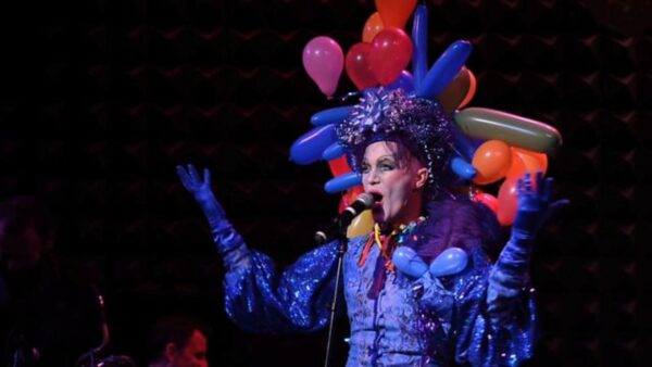 A person in a blue dress with a balloon headdress stands in front of a microphone with their mouth open