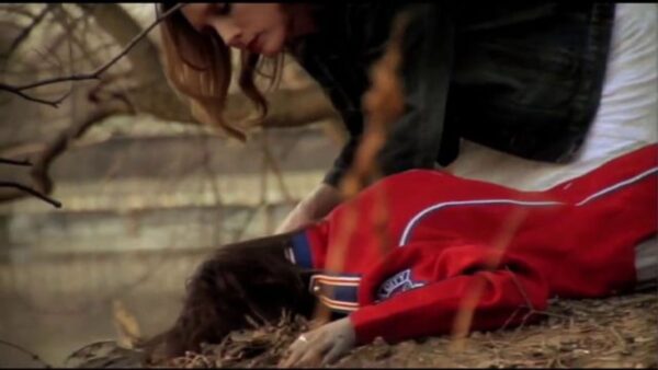 A person leans over a person with a red marching band coat lying face down in the middle of dead grass