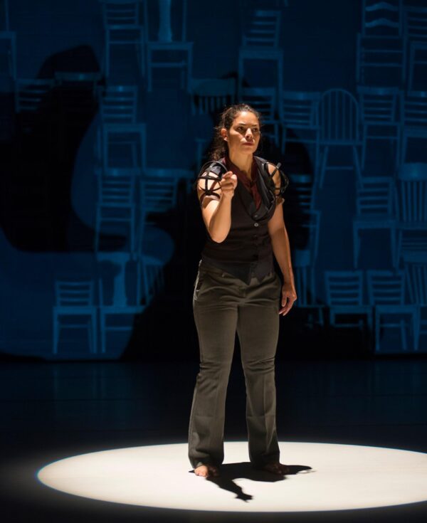 A singular, spotlit performer in all black points toward the audience in front of a background of stacked chairs