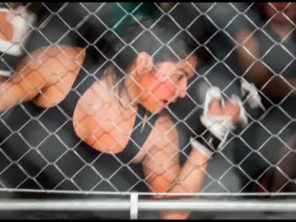 A boxer poses behind a chain link fence, arms flexed outwards on each side of her body