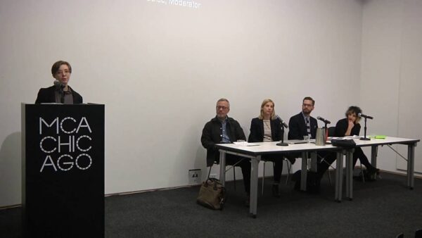 One person stands behind a black podium with the MCA Chicago logo on it as four people sitting at a table look on