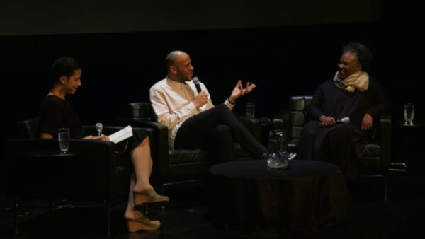 Three people sit on a dark stage looking at the person in the middle who holds a microphone up to their mouth