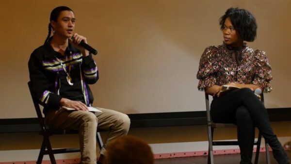 Two people sit on a stage in folding chairs as one holds a microphone up to their mouth