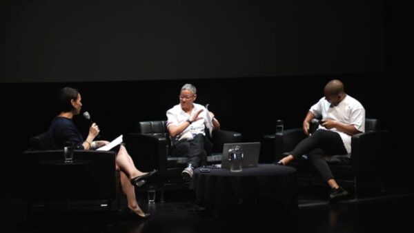 Three people sit in chairs situated around three sides of a coffee table on a black stage