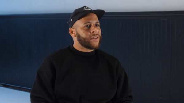 A male-presenting person from the waist up in a black sweatshirt and baseball cap with a close-cut beard in a room that has a long horizontal strip of gray on the wall