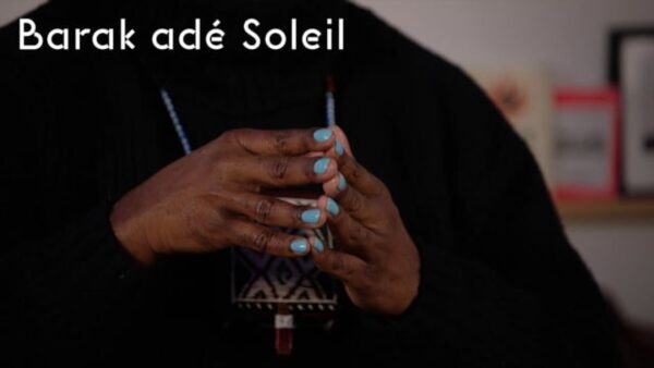 Close up of dark brown hands with robin's egg blue finger nails tented in front of somebody's chest wearing a black shirt. White text at the top of the image reads Barak ade Soleil.