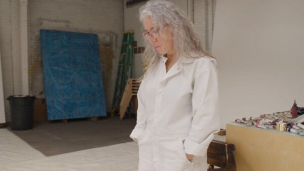 A female-presenting person with shoulder-length grey hair looks down at the floor in a studio space