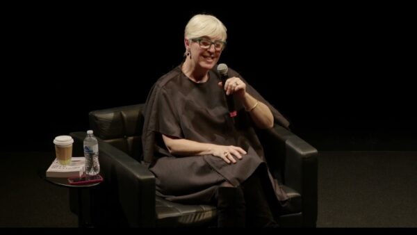 A woman with alabaster skin, dressed in all black with platinum blond hair, sits in a black chair on a black stage holding a microphone. A small table holds a coffee, water bottle, and book.