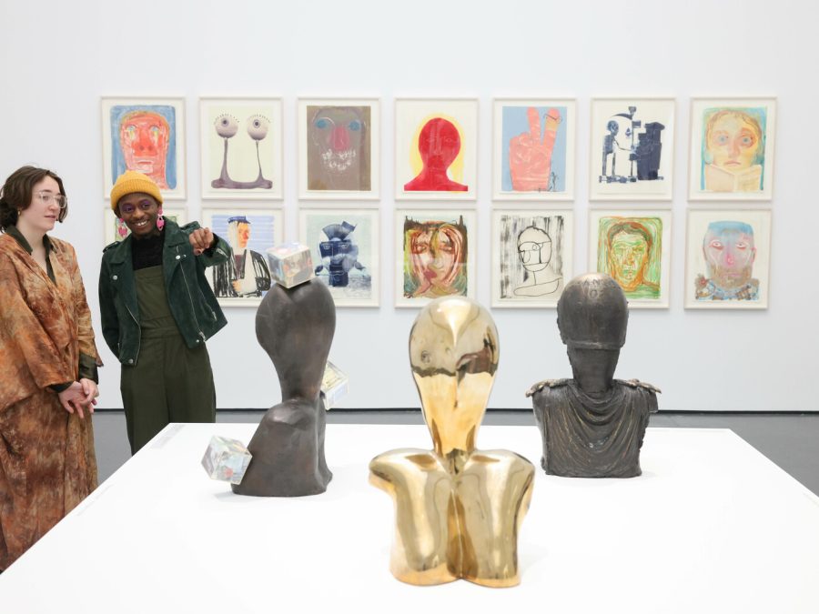 Two people look at a set of three sculptures in a gallery space with a series of drawings on the wall behind them.
