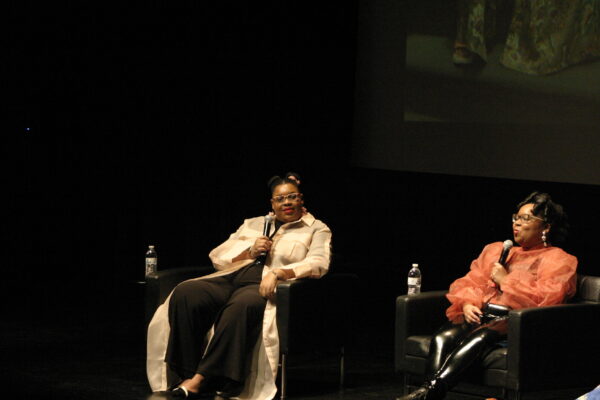 Two brown-skinned female presenting people sit on a darkened stage holding microphones