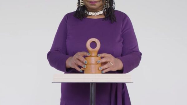 The torso of a person with light brown skin wearing a purple shirt and a puka shell necklace rests their hands on either side of a small clay sculpture with a looped top handle.