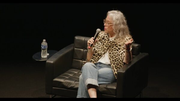 A female-presenting person with light skin dressed in a leopard print shirt and jeans sits on a black stage in a black chair holding a microphone. A small table next to them has a water bottle on it.