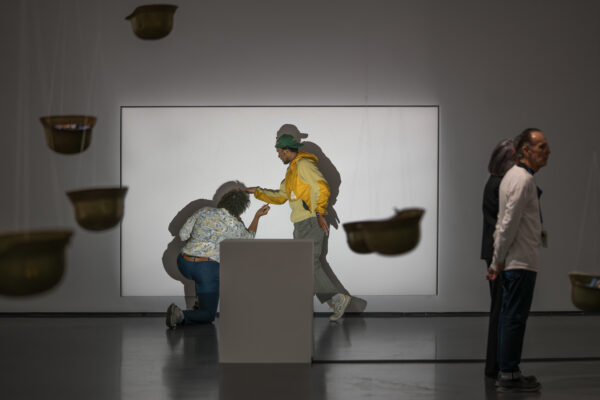 A person traces another person's shadow posed in a walking motion on a white wall in a room filled with hanging helmets