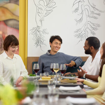 Four adults sit at a dining table with wine glasses and plates in front of them as they talk.