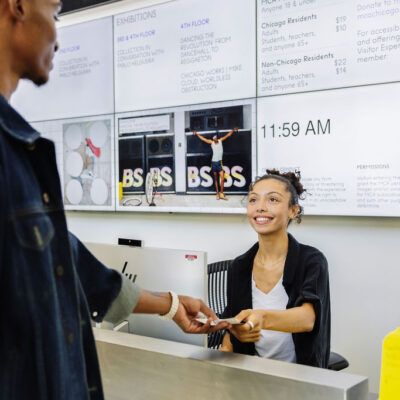 A young woman behind a computer and sitting in a desk chair smiles as she hands two young men tickets.