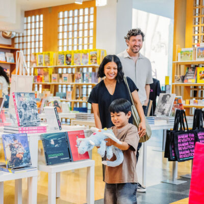 A young boy walks in front of a woman and man, smiling, as he holds a stuffed animal.