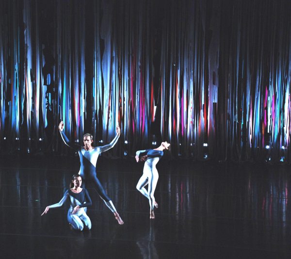 Three dancers on a stage with a shimmery curtain lit up in blues and purples behind them