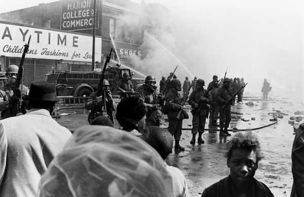 Archival black and white image of a group of people holding guns in gas masks. In front of them a small Black child looks directly at the camera while standing near other people.