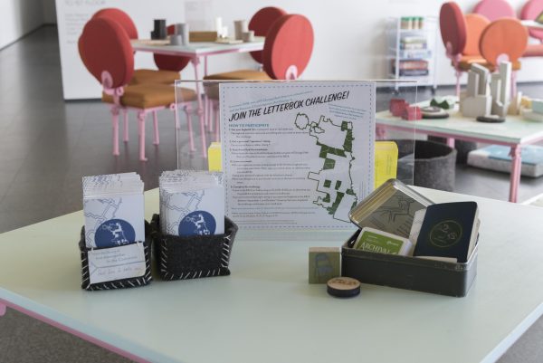 A table with an instruction sign, stamps, and paper on in in a room filled with orange chairs around tables