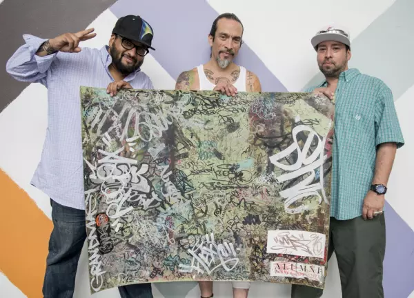 Three people hold a tapestry with graffiti on it