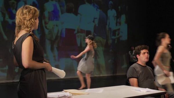 Five people on a stage in various positions not interacting with each other. A projection of people is on a screen behind them.
