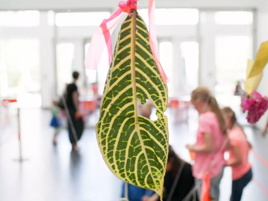 A large green leaf with yellow stripes, tied with pink ribbons, hangs in the foreground. In the background, a group of people are gathered, engaging in an art or craft activity inside a bright and spacious room.