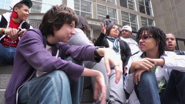 A group of kids sits on the steps outside of a grey building filled with windows