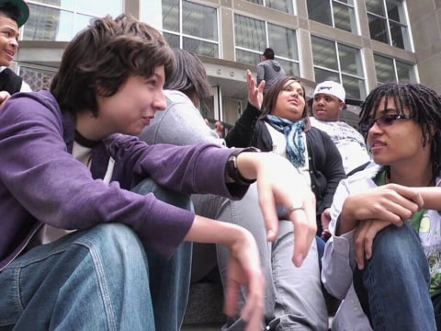 A group of kids sits on the steps outside of a grey building filled with windows
