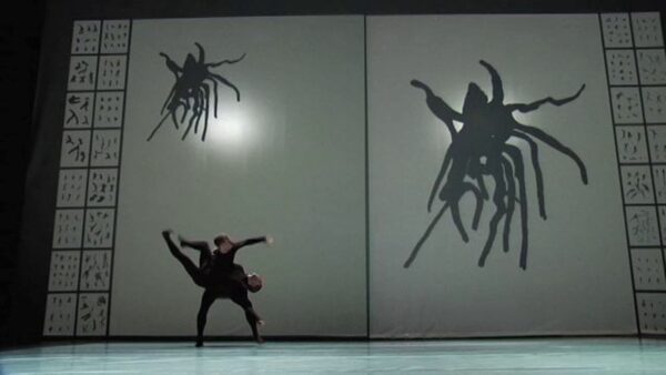 Two entwined dancers on a stage in front of a backdrop of a leggy creature which appears small on the upper-left corner of the backdrop and large on the right side.