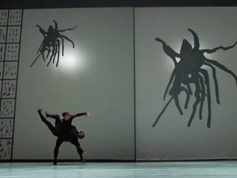 Two entwined dancers on a stage in front of a backdrop of a leggy creature which appears small on the upper-left corner of the backdrop and large on the right side.