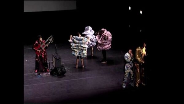 Two singers and a double guitarist perform on a stage with three people in full, layered fabric costumes behind them