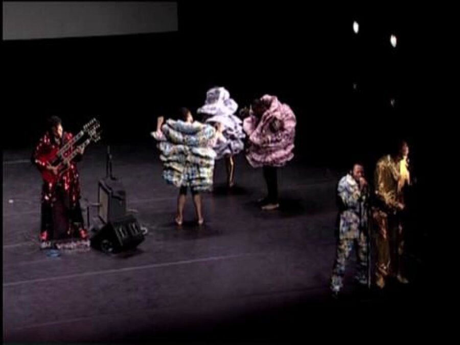 Two singers and a double guitarist perform on a stage with three people in full, layered fabric costumes behind them
