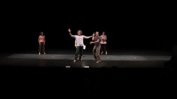 Four people at various depths from the audience on a stage lit only by one light. The person in the middle stands with their arms spread wide.