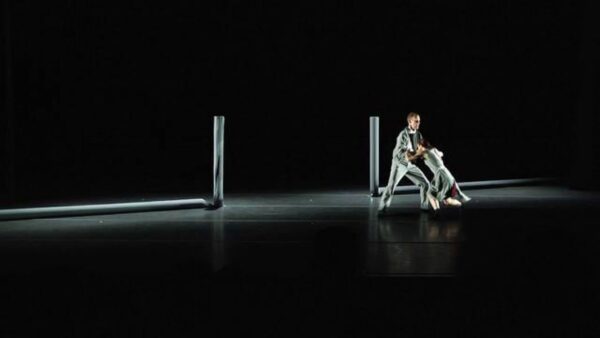 Two dancers push against each other on a stage with a simple set of two posts