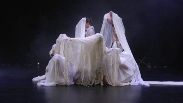 People wrapped in a large white sheet on a black stage
