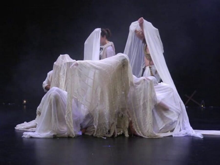 People wrapped in a large white sheet on a black stage