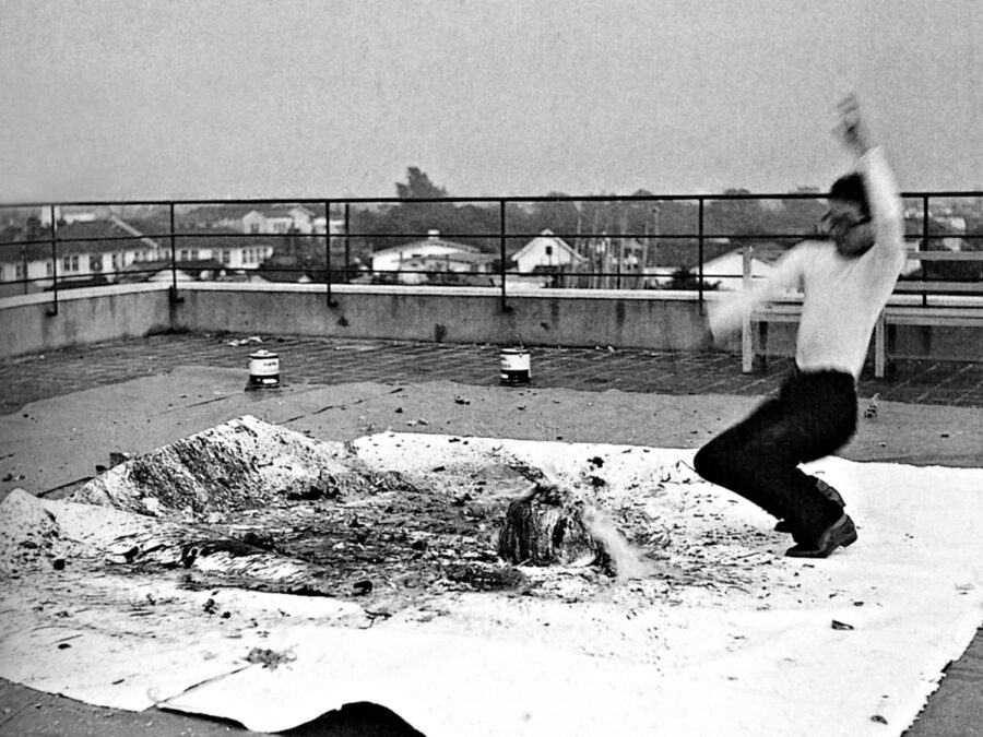 Black and white photograph of a person standing in an active position on a large white square covered in objects and splattered liquid