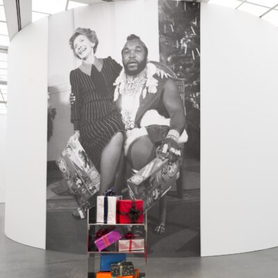 A larger than life black-and-white image of Nancy Reagan sitting on Mr. T's lap on a white wall behind a cart of wrapped presents