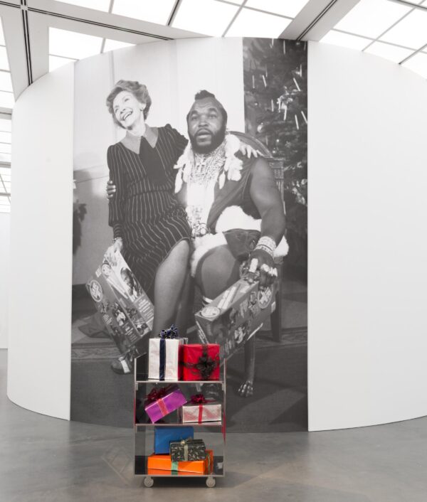 A larger than life black-and-white image of Nancy Reagan sitting on Mr. T's lap on a white wall behind a cart of wrapped presents