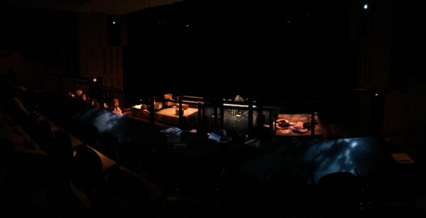 A person bends over a table in a darkened theater with a few people in the audience and pillows on the stage