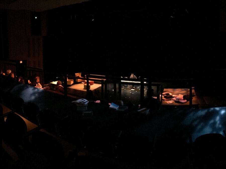 A person bends over a table in a darkened theater with a few people in the audience and pillows on the stage