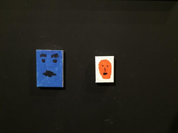 Two simplistic paintings of faces, one blue with black features, one orange with black features against a white background, hanging on a black wall