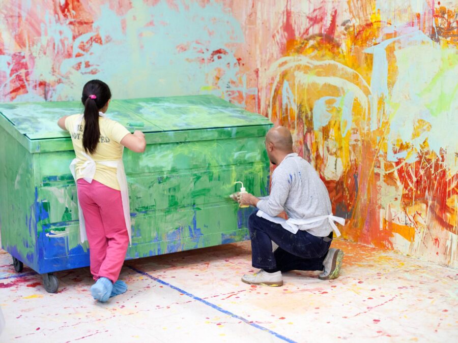 Two people paint a dumpster green in a space with colorful, haphazardly painted walls in pinks, blues, reds, and yellows