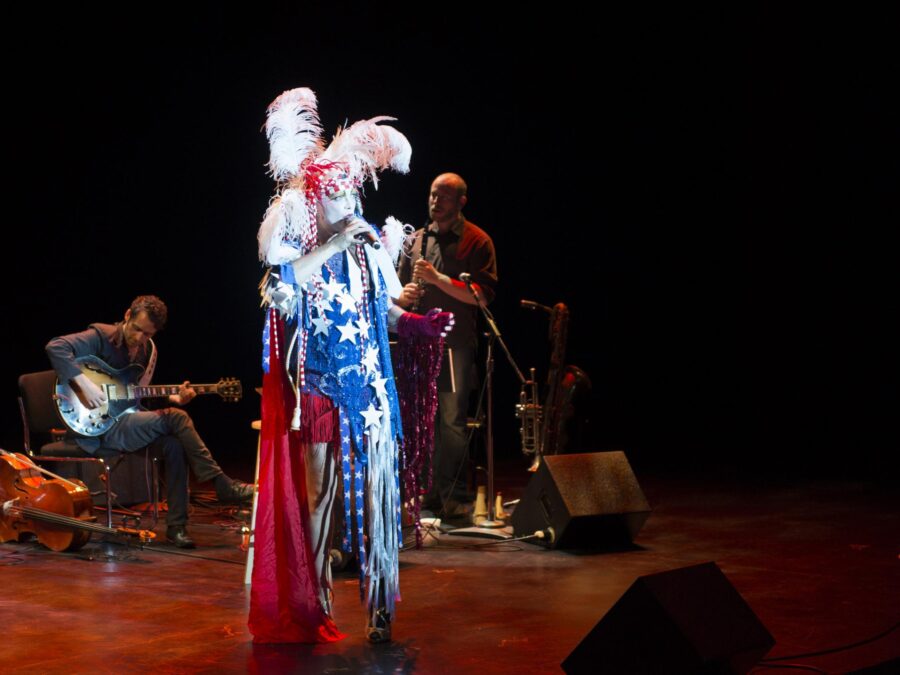 A person wearing a costume with a cape in red, white, and blue, and a feathered headdress sings on stage in the spotlight.