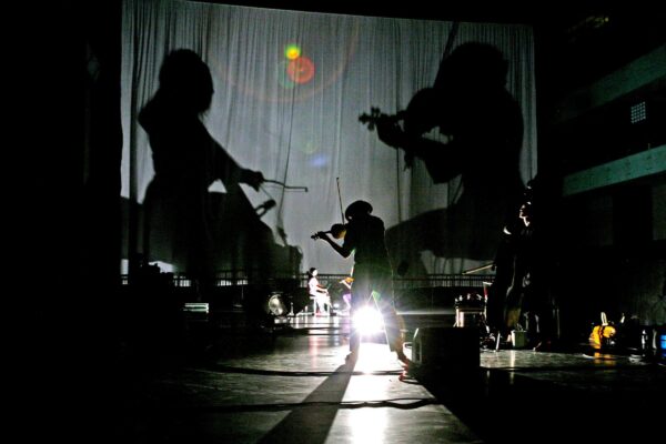 The shadows of two musicians are cast on a curtain on a dark stage where instruments and one person playing the violin are visible