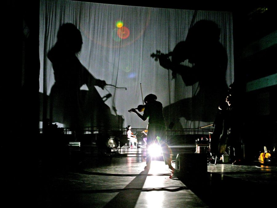 The shadows of two musicians are cast on a curtain on a dark stage where instruments and one person playing the violin are visible